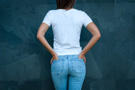 White mockup t-shirt for print design. Rear view or back view of woman in blank white t-shirt and blue jeans on the grey backgroundの写真素材