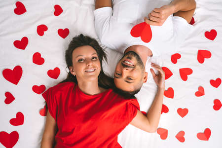 Top view of attractive couple in love lying on the bed with red paper heartsの写真素材