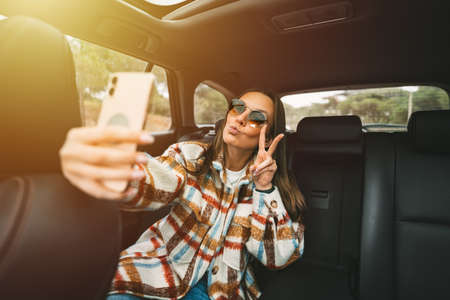 Stylish young woman sitting on back seat in the car and making selfie with smartphone. Woman taking picture with phone while sitting in taxiの写真素材