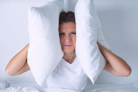 Young woman disturbed by noisy neighbours. Woman struggling from noise in bed and covering ears with...