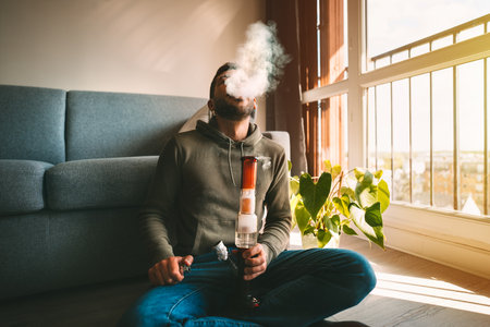 Man smoking bong and exhaling the smoke at home. Man smoking pot, medical marijuana or cannabis from...