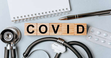 Wooden block form the word COVID with stethoscope, pills, notepad, pen on the doctor's desktop. Medical concept.の写真素材
