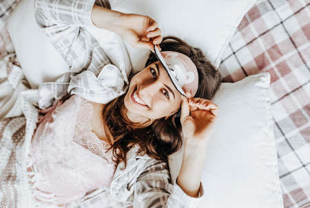 A sleepy brunette girl lies in bed with a blindfold waking up in the morning. Cozy home morning in cute pajamas. Have a good time, relax and dream. Healthy sleep.の写真素材