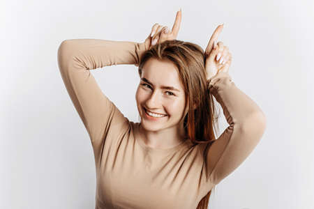 Young beautiful girl smiles and makes horns with fingers gesture on a white isolated background. A woman fooling around and grimacing. Place for advertising. Positive brunette in a beige jumper.の写真素材
