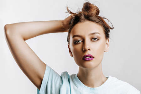 A beautiful brunette girl holds her hair and makes her own hairstyle. Young model with long hair, fashion makeup with purple lips on an gray background. The woman seriously looking at the camera.の写真素材