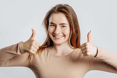 Beautiful young girl smiling and shows thumbs up gesture with two hands on a white isolated background. A woman points to an idea, a place for advertising. Positive brunette in a beige jumper.の写真素材