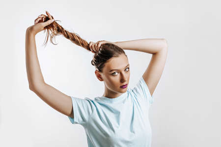 A beautiful brunette girl holds her hair and makes her own hairstyle. Young model with long hair, fashion makeup with purple lips on an gray background. The woman seriously looking at the camera.の写真素材