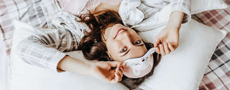 A sleepy brunette girl lies in bed with a blindfold waking up in the morning. Cozy home morning in cute pajamas. Have a good time, relax and dream. Healthy sleep.の写真素材