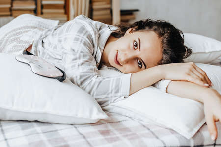 A sleepy brunette girl lies in bed with a blindfold waking up in the morning. Cozy home morning in cute pajamas. Have a good time, relax and dream. Healthy sleep.の写真素材