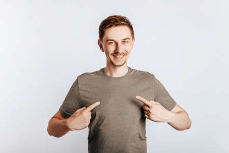 An attractive man points with both hands to himself. Handsome guy proposes and smiles. Kind young businessman over isolated gray background. Place for advertising.の写真素材