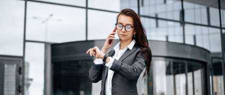 A business woman checks the time and talking on the phone in the city during a working day waiting for a meeting. Discipline and timing. An employee goes towards a corporate meeting.の写真素材