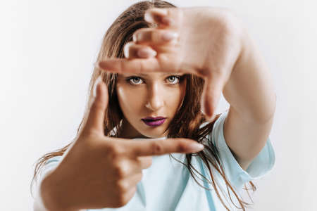 Beautiful young girl makes a frame with her hands on an isolated gray background. The woman with fashion make-up with purple lips seriously looking at the camera. Place for advertising.の写真素材