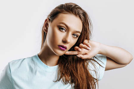 Beautiful young brunette girl with green eyes with long hair with fashion makeup with purple lips on an isolated gray background. The woman holds hand to face and seriously looking at the camera.の写真素材