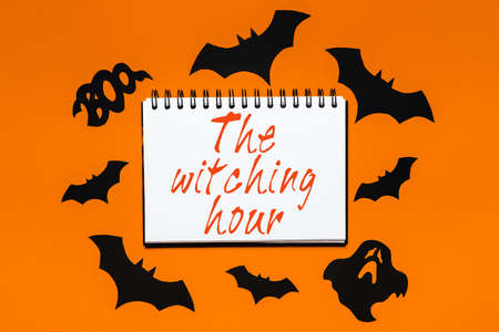 Happy halloween holiday concept. Notepad with text The witching hour on white and orange background with bats, pumpkins and ghostsの写真素材