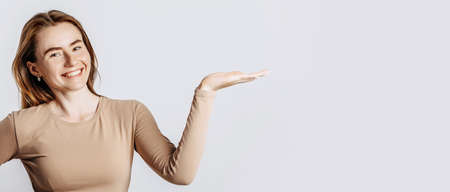 Beautiful young girl smiling and pointing her hand to the side at on a white isolated background. A woman points to an idea, a place for advertising. Positive brunette in a beige jumper.の写真素材