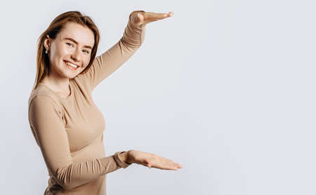 Young beautiful girl smiles and shows big size measure with her hand gesture on a white isolated background. A woman points to an idea, a place for advertising. Positive brunette in a beige jumper.の写真素材
