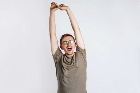 Good morning. The sleepy man yawns, pulls his arms up and stretches. Boredom and sleepiness. Young handsome man with glasses a brunette beard posing on gray background. Place for advertising.の写真素材