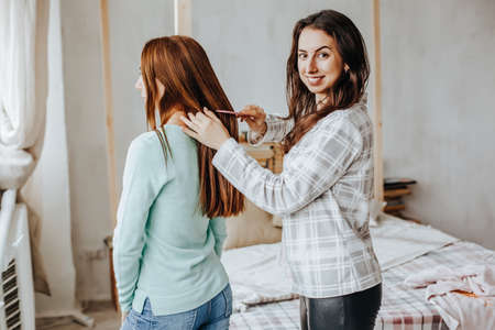 Two girls braid their hair at the window. Woman makes a braid to her friend. Hair weaving hairstyles. Girlfriend braids her hands with ringlets. Hair careの写真素材
