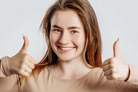 Beautiful young girl smiling and shows thumbs up gesture with two hands on a white isolated background. A woman points to an idea, a place for advertising. Positive brunette in a beige jumper.の写真素材