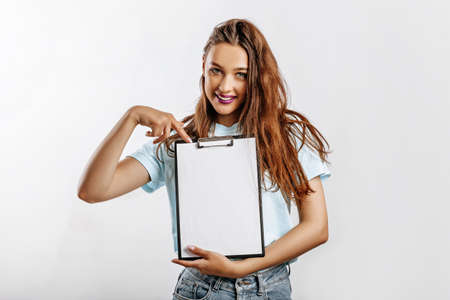 Young beautiful girl smiling and holding a clip board over gray background. The student points her finger at the sheet. Business concept. Woman with fashion make-up purple lips. Place for advertising.の写真素材
