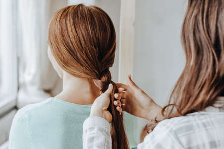 Two girls braid their hair at the window. Woman makes a braid to her friend. Hair weaving hairstyles. Girlfriend braids her hands with ringlets. Hair careの写真素材
