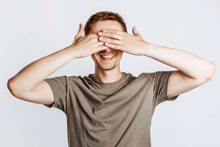 Handsome young man closes his eyes with two hands and smiles. Brunette with a beard in a green shirt posing on a gray background. Place for advertising. I do not see.の写真素材