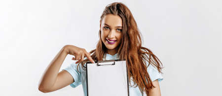 Young beautiful girl smiling and holding a clip board over gray background. The student points her finger at the sheet. Business concept. Woman with fashion make-up purple lips. Place for advertising.の写真素材