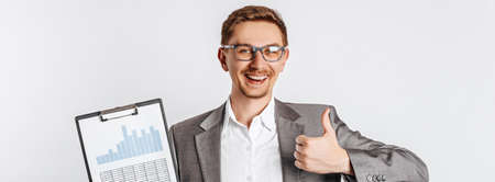 Young handsome brunette man in glasses in a suit smiles and shows thumbs up with business documents and graphs on white isolated backgroundの写真素材