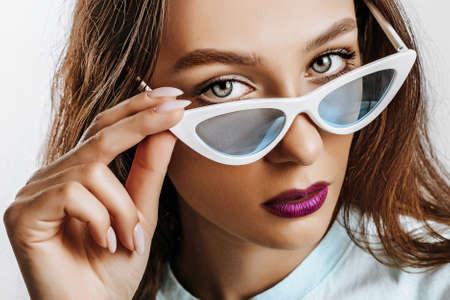 Beautiful young brunette girl with green eyes with fashion makeup with purple lips. The woman in blue sunglasses seriously looking at the camera. Beauty concept.の写真素材