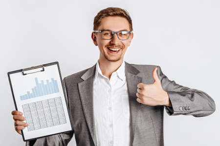 Young handsome brunette man in glasses in a suit smiles and shows thumbs up with business documents and graphs on white isolated backgroundの写真素材