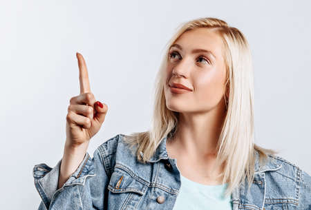 Portrait of a smiling girl pointing finger up at copyspace isolated on a white background. A woman points to an idea, a place for advertising. Positive blonde.の写真素材