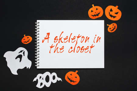 Happy halloween holiday concept. Notepad with text A skeleton in the closet on black background with bats, pumpkins and ghostsの写真素材