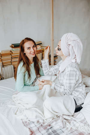 Two girls make masks and patches for the beauty of face and hair. Women take care of youthful skin. Girlfriends laugh, make faces and do beauty treatments at home with towels on their heads.の写真素材