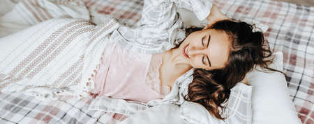 A sleepy brunette girl lies in bed with a blindfold waking up in the morning. Cozy home morning in cute pajamas. Have a good time, relax and dream. Healthy sleep.の写真素材