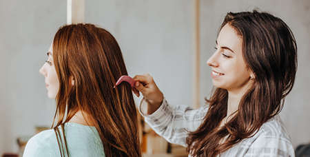 Two girls braid their hair at the window. Woman makes a braid to her friend. Hair weaving...