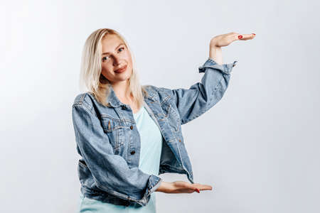 Smiling blonde girl shows big size with her hands on a gray background in the studio. A woman in casual clothes is happy with the measure. Place for advertising.の写真素材