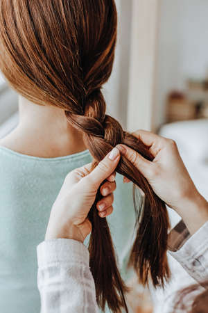 Two girls braid their hair at the window. Woman makes a braid to her friend. Hair weaving hairstyles. Girlfriend braids her hands with ringlets. Hair careの写真素材
