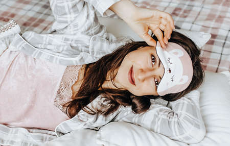 A sleepy brunette girl lies in bed with a blindfold waking up in the morning. Cozy home morning in cute pajamas. Have a good time, relax and dream. Healthy sleep.の写真素材