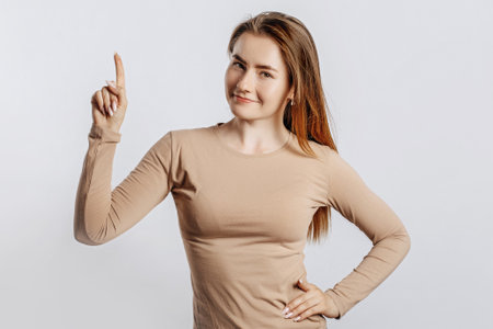 Beautiful young girl smiling and pointing her finger to the side at on a white isolated background. A woman points to an idea, a place for advertising. Positive brunette in a beige jumper.の写真素材