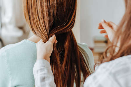 Two girls braid their hair at the window. Woman makes a braid to her friend. Hair weaving hairstyles. Girlfriend braids her hands with ringlets. Hair careの写真素材