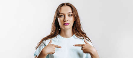 A beautiful young girl smiles and points her fingers at herself on a white isolated gray background. Place for advertising. Positive woman with fashion make-up with purple lips.の写真素材