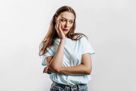Beautiful young brunette girl with green eyes with long hair with fashion makeup with purple lips on an isolated gray background. The woman holds hand to face and looks to the side seriously and sadlyの写真素材