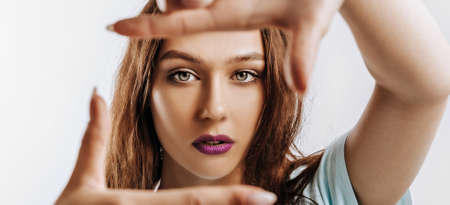 Beautiful young girl makes a frame with her hands on an isolated gray background. The woman with fashion make-up with purple lips seriously looking at the camera. Place for advertising.の写真素材