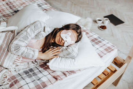 A sleepy brunette girl lies in bed with a blindfold waking up in the morning. Cozy home morning in cute pajamas. Have a good time, relax and dream. Healthy sleep.の写真素材