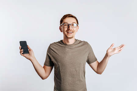 Young handsome man holding a smartphone and smiling. The guy shows his hands and raises his shoulders. Student, worker posing for photo isolated on gray background. Place for advertising.の写真素材