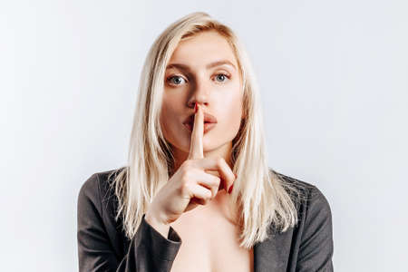 Beautiful young serious girl showing hush gesture, keep secret on gray isolated backgroundの写真素材