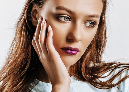 Beautiful young brunette girl with green eyes with long hair with fashion makeup with purple lips on an isolated gray background. The woman holds hand to face and looks to the side seriously and sadlyの写真素材