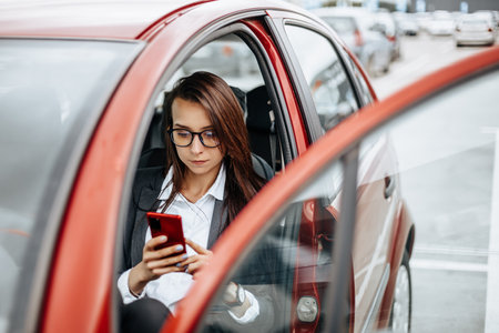 The girl in the car behind the wheel looks at the notification phone and reads the message. A woman in a parking lot sits in a auto.の写真素材