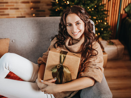 Beautiful cheerful happy young girl with christmas gifts on sofa on the background of a new year tree at homeの写真素材