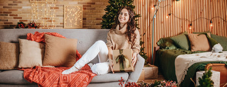 Beautiful cheerful happy young girl with christmas gifts on sofa on the background of a new year tree at homeの写真素材